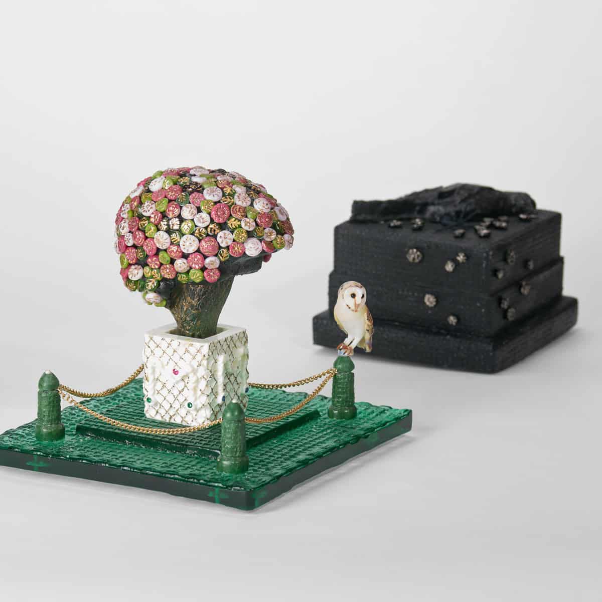 Pink and white floral glass tree with owl figurine on a green base, black textured block in background.
